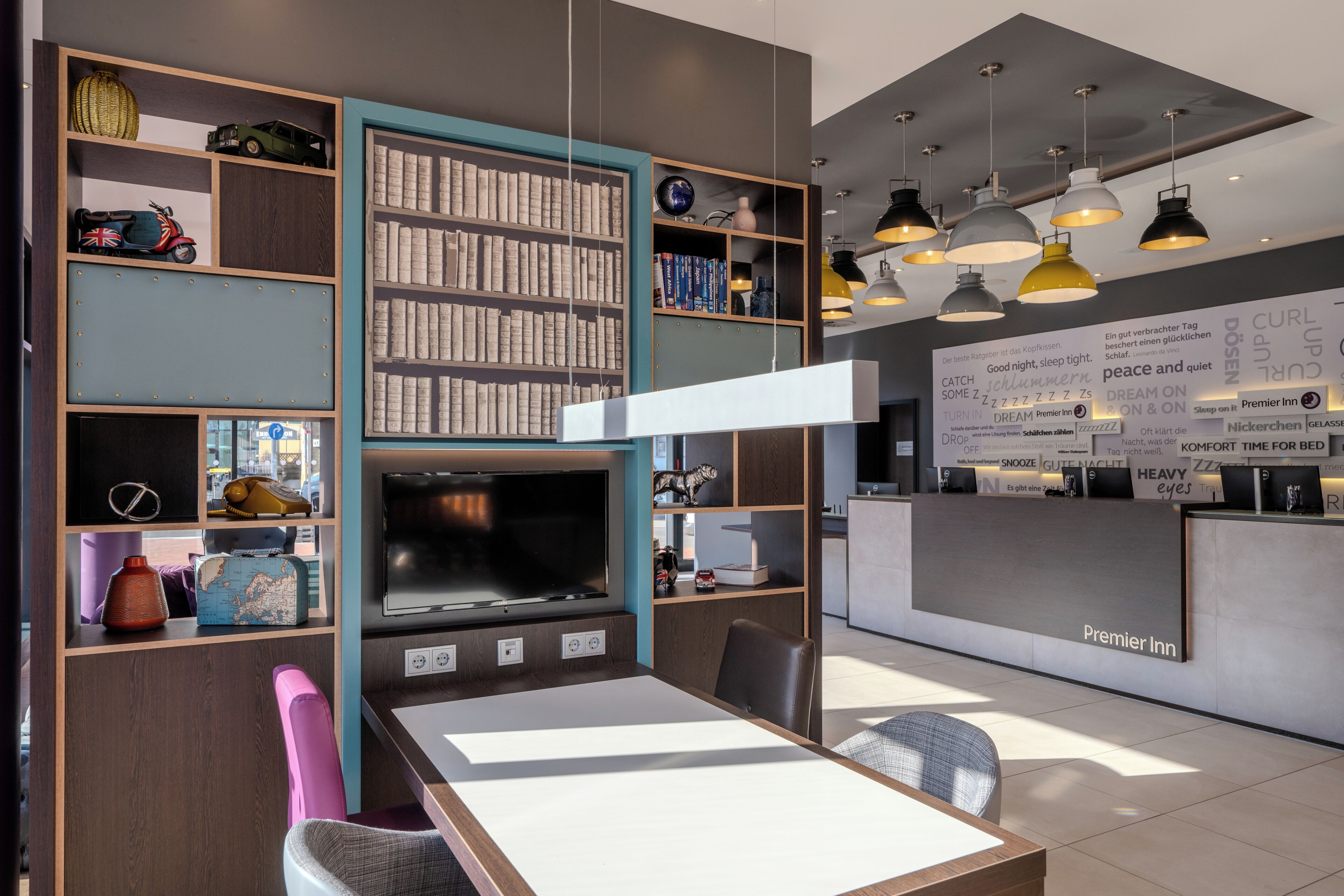 A modern hotel room with a large flat-screen TV, a dining area with a table and chairs, a bookshelf with various items, and a reception desk with a sign.