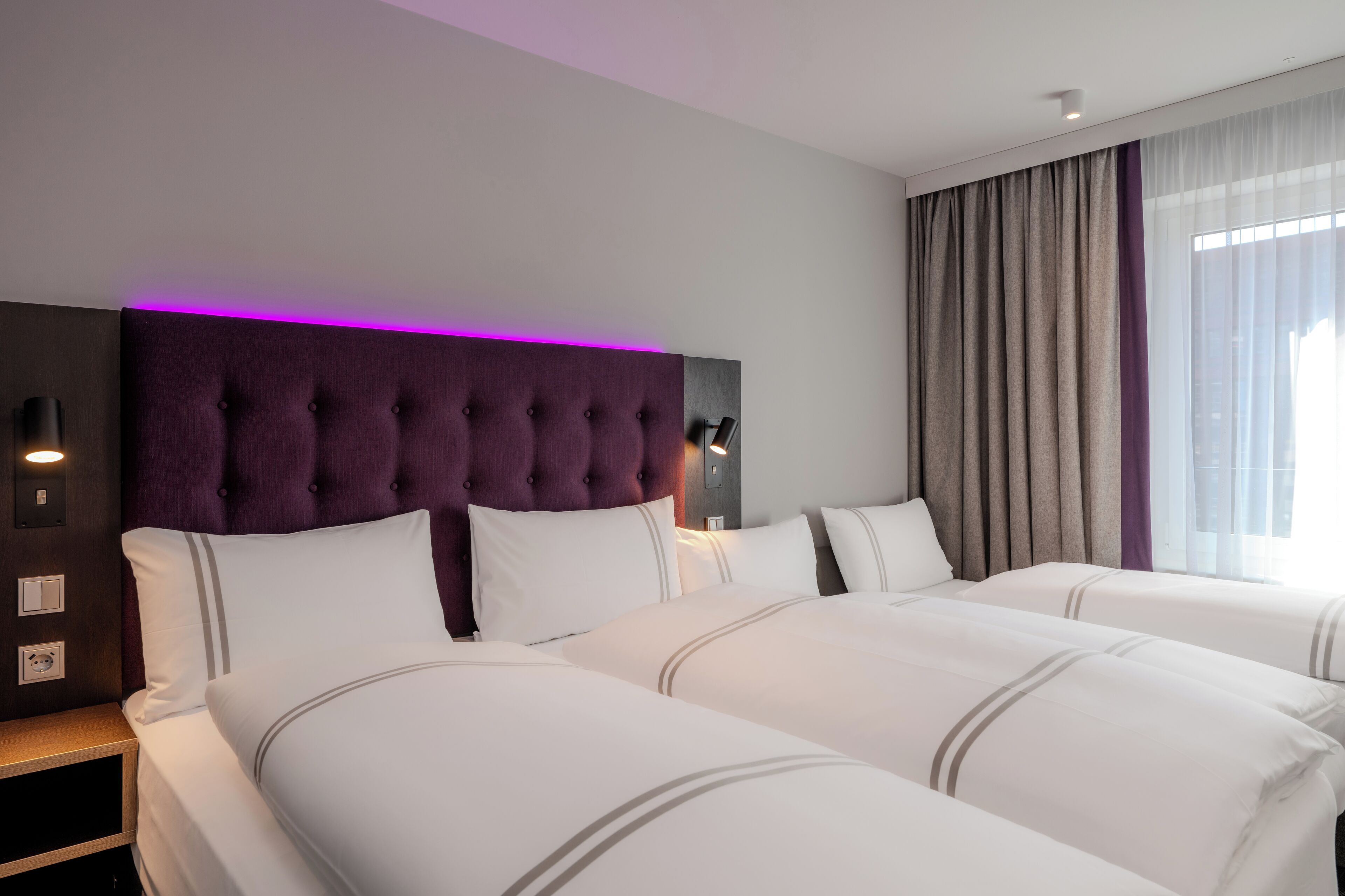 A hotel room with a tufted headboard, two beds with white bedding, a nightstand, a wall-mounted lamp, and a window with curtains.