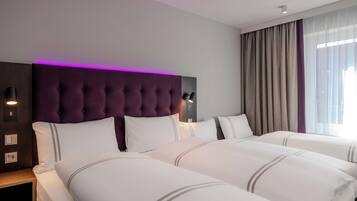 A hotel room with a tufted headboard, two beds with white bedding, a nightstand, a wall-mounted lamp, and a window with curtains.