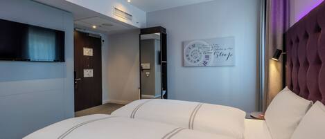 Une chambre d’hôtel avec un grand lit, un téléviseur à écran plat, un miroir, une horloge affichant une citation inspirante et une tête de lit violette.