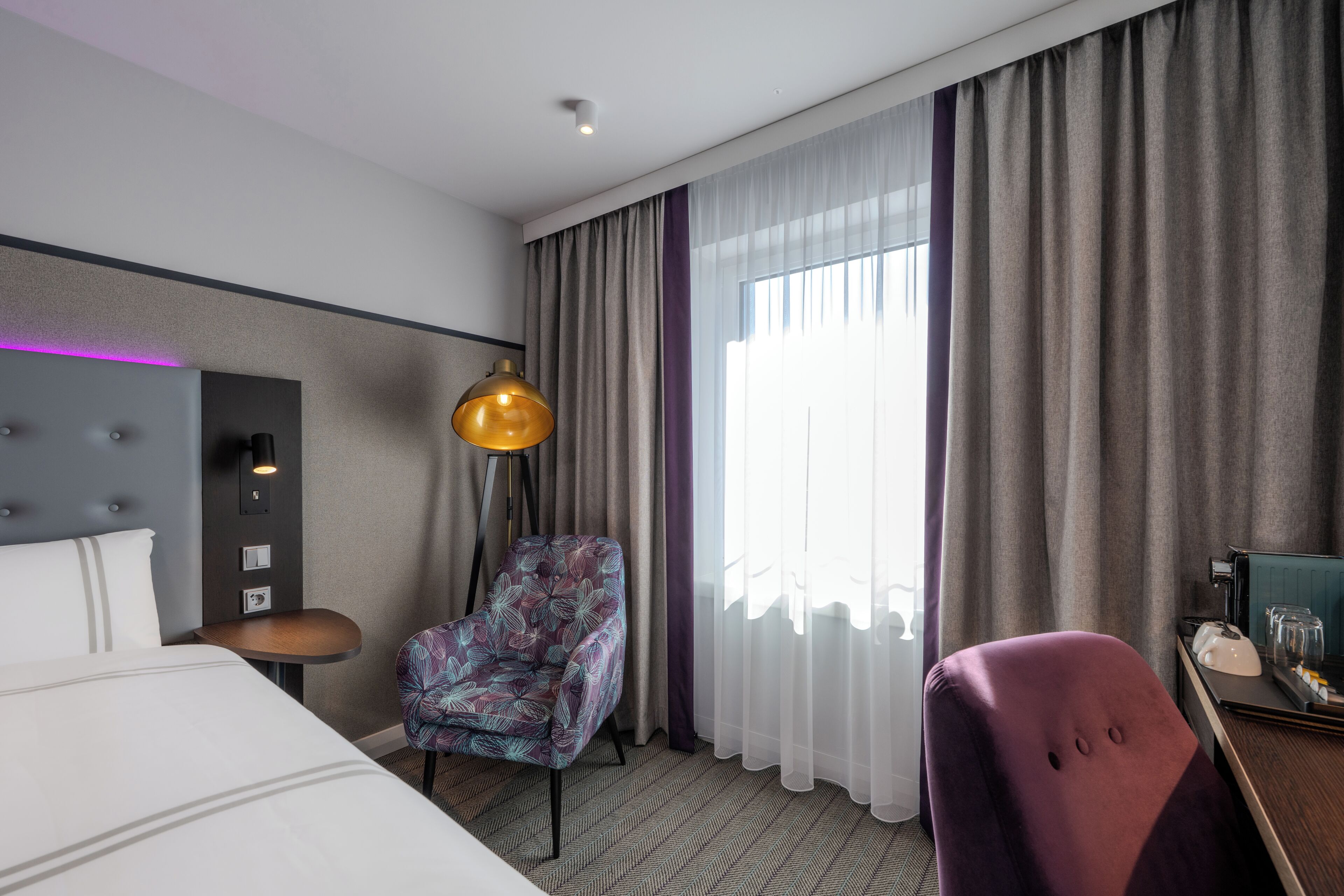 Ein Hotelzimmer mit Bett, Stuhl, Schreibtisch, Lampe und einem fensterbepflegten Fenster.