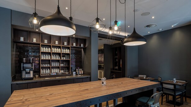 A modern bar area with a long wooden counter, multiple seating options, and pendant lighting. - Premier Inn Hannover City Centre (Hannover)