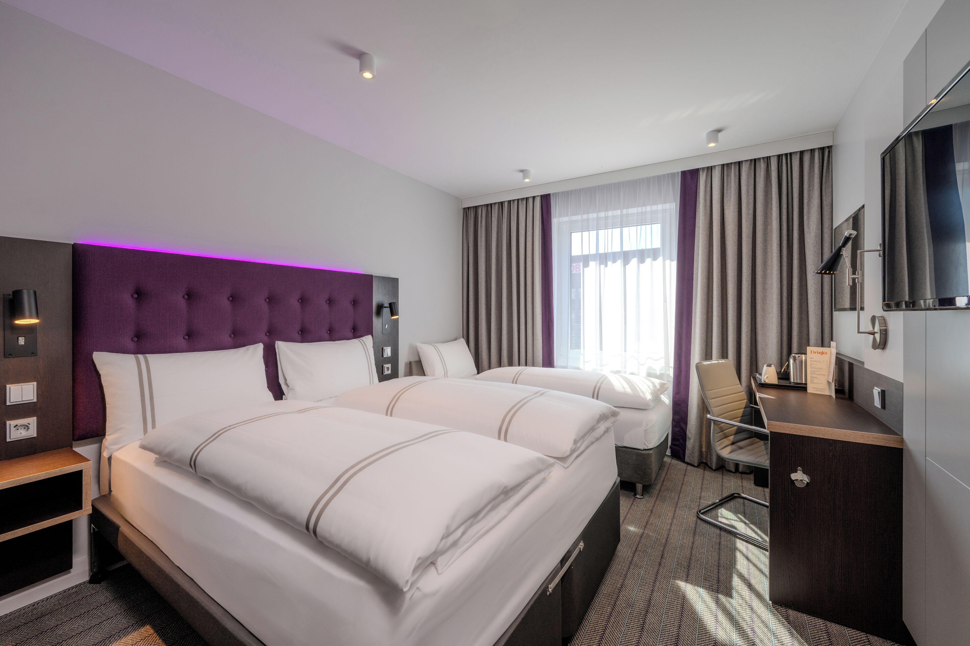 A hotel room with two beds, a desk, a TV, and a large window with curtains.