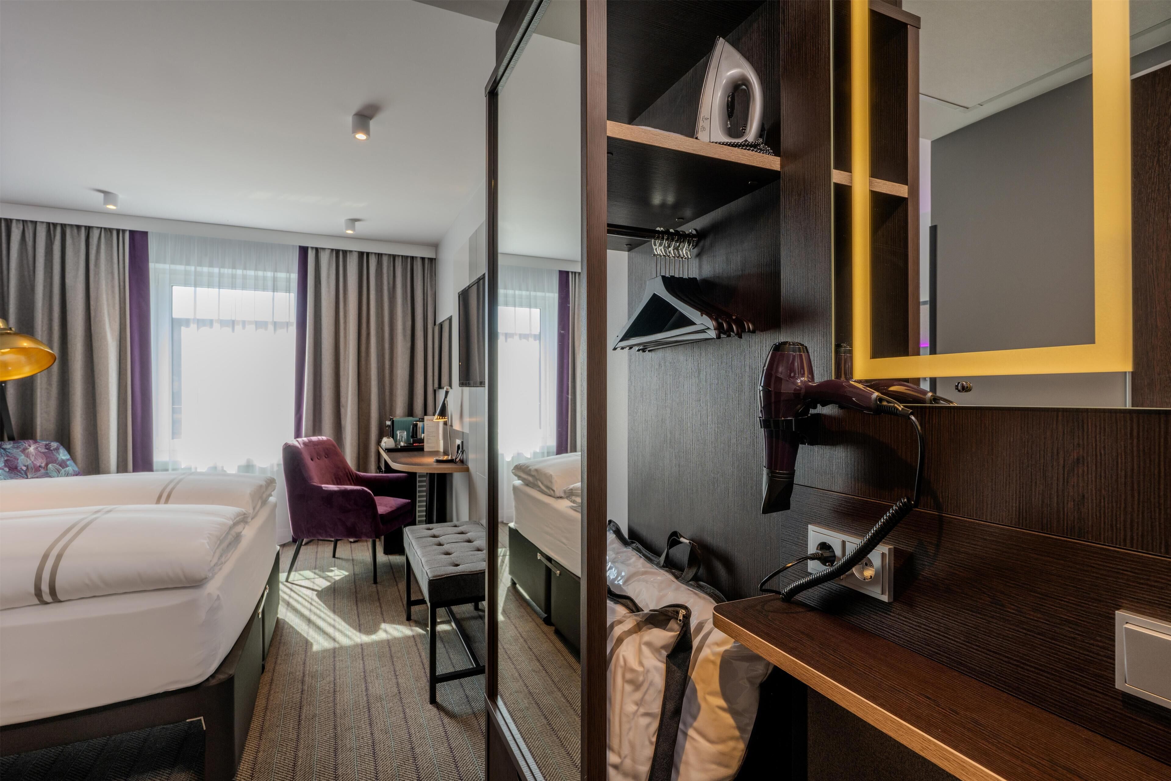A hotel room with two beds, a desk with a chair, a mirror, a wardrobe, and a suitcase.