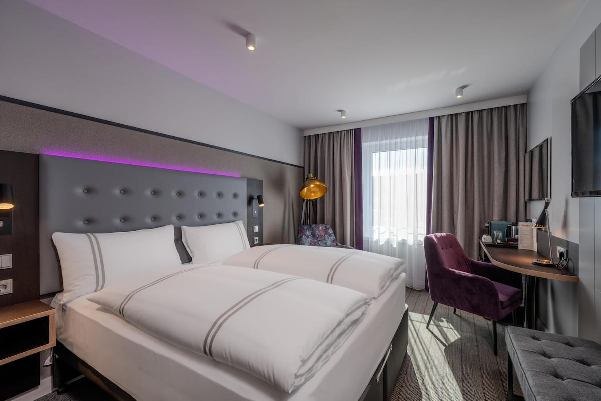 Une chambre d’hôtel moderne dotée d’un grand lit, d’un bureau avec une chaise, d’une petite table et d’une grande fenêtre avec des rideaux.