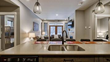 A modern kitchen with a central island, built-in dishwasher, and wooden cabinetry. There is a dining area with a table and chairs, and a living room with a sofa and a fireplace.