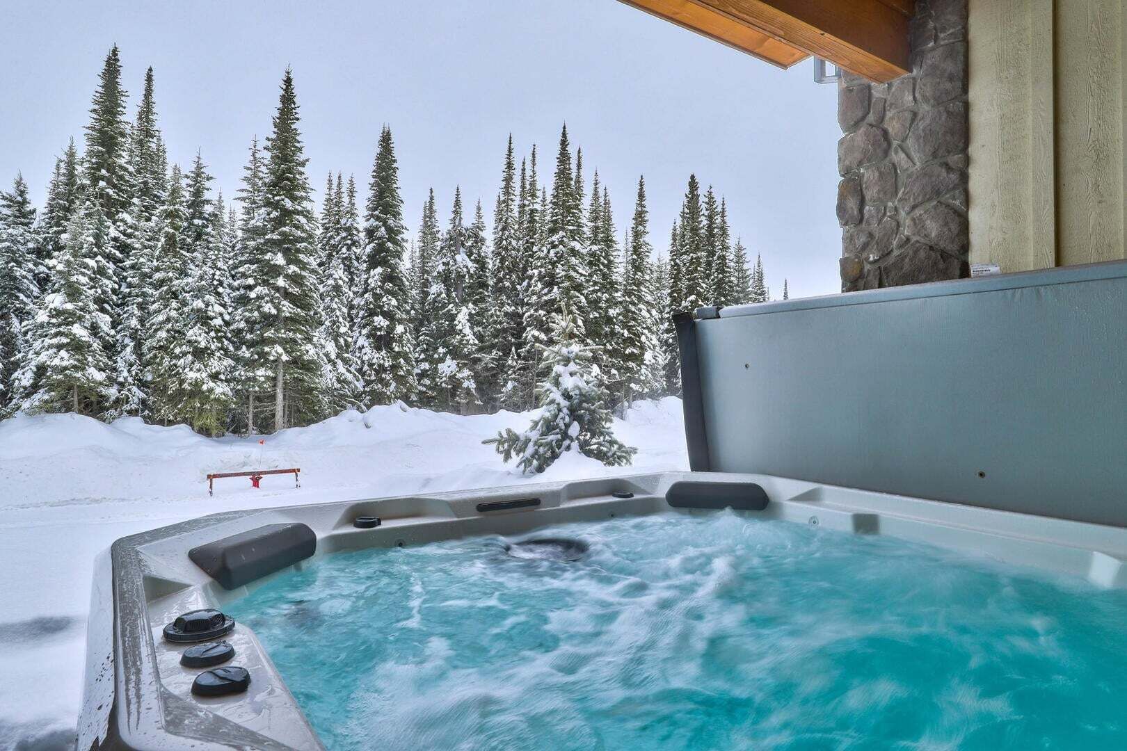 A hot tub with a view of snow-covered trees and a stone wall.