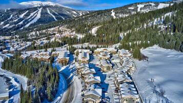 A snow-covered resort with multiple chalet-style buildings arranged along a winding road, surrounded by dense forests and snow-covered mountains.