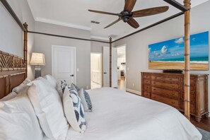 A bedroom with a large bed, a ceiling fan, a wooden dresser, and a painting of a beach scene. - Summerwoods 1903 2 Bedroom Condo (Fernandina Beach)