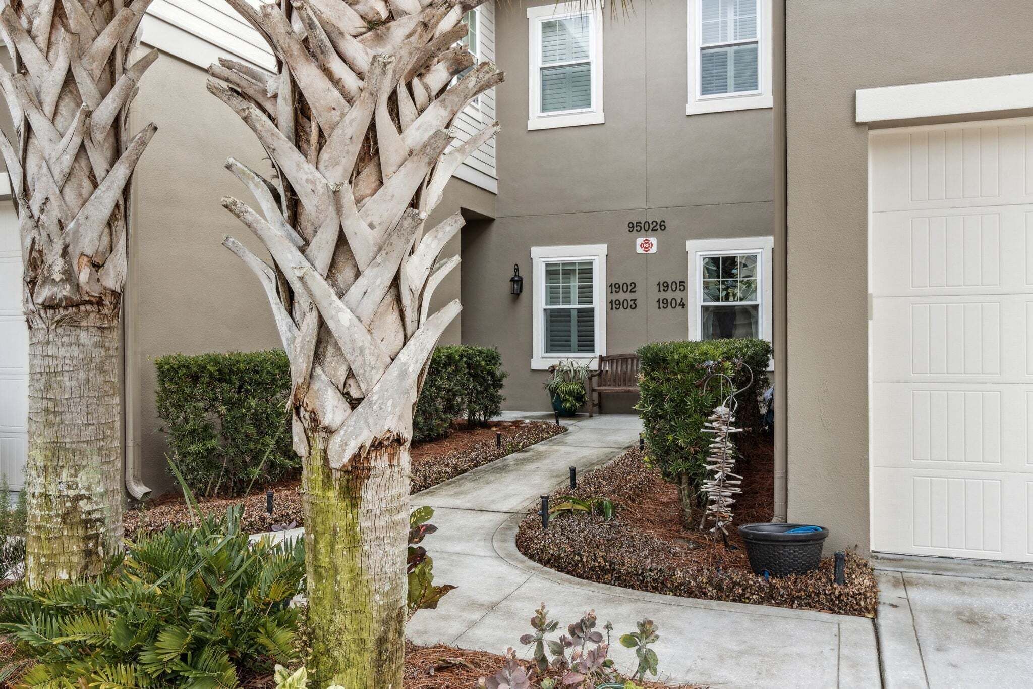 A residential building with a driveway leading to a garage, surrounded by a landscaped garden with various plants and a palm tree.