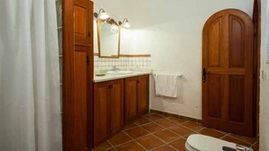 Une salle de bain avec des meubles en bois, un lavabo blanc, un miroir avec éclairage, une serviette accrochée au mur et une porte en bois.