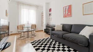Una sala de estar moderna con un sofá gris, una mesa de comedor de vidrio y sillas, un tapete blanco y negro, y un televisor.