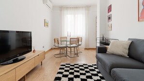 Una sala moderna con una televisión de pantalla plana, un sofá gris, una mesa de comedor con sillas y un tapete de cuadros.