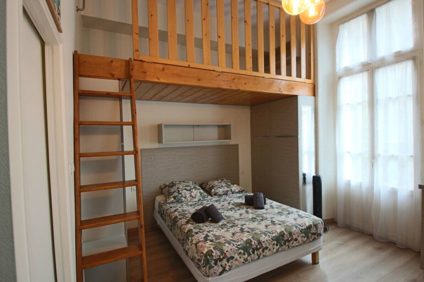 Een slaapkamer met een stapelbed, een houten trap, een bed met bloemenbeddengoed en een raam met dunne gordijnen.