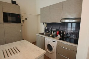 A compact kitchen with a microwave, washing machine, and a cityscape view through the window. - Studio 25m², en hyper centre-ville (Aix-les-Bains)
