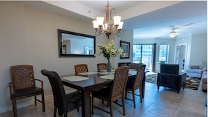 A dining area with a wooden table, six chairs, a chandelier, and a vase with flowers. - Ocean View 3BR Condo – Unit 208 Royale Palms (Myrtle Beach)