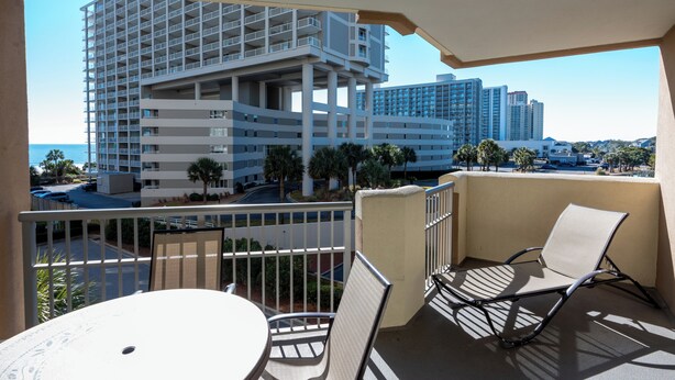 A balcony with a table and chairs, overlooking a parking area and a building with multiple floors and balconies. - Ocean View 3BR Condo – Unit 208 Royale Palms (Myrtle Beach)