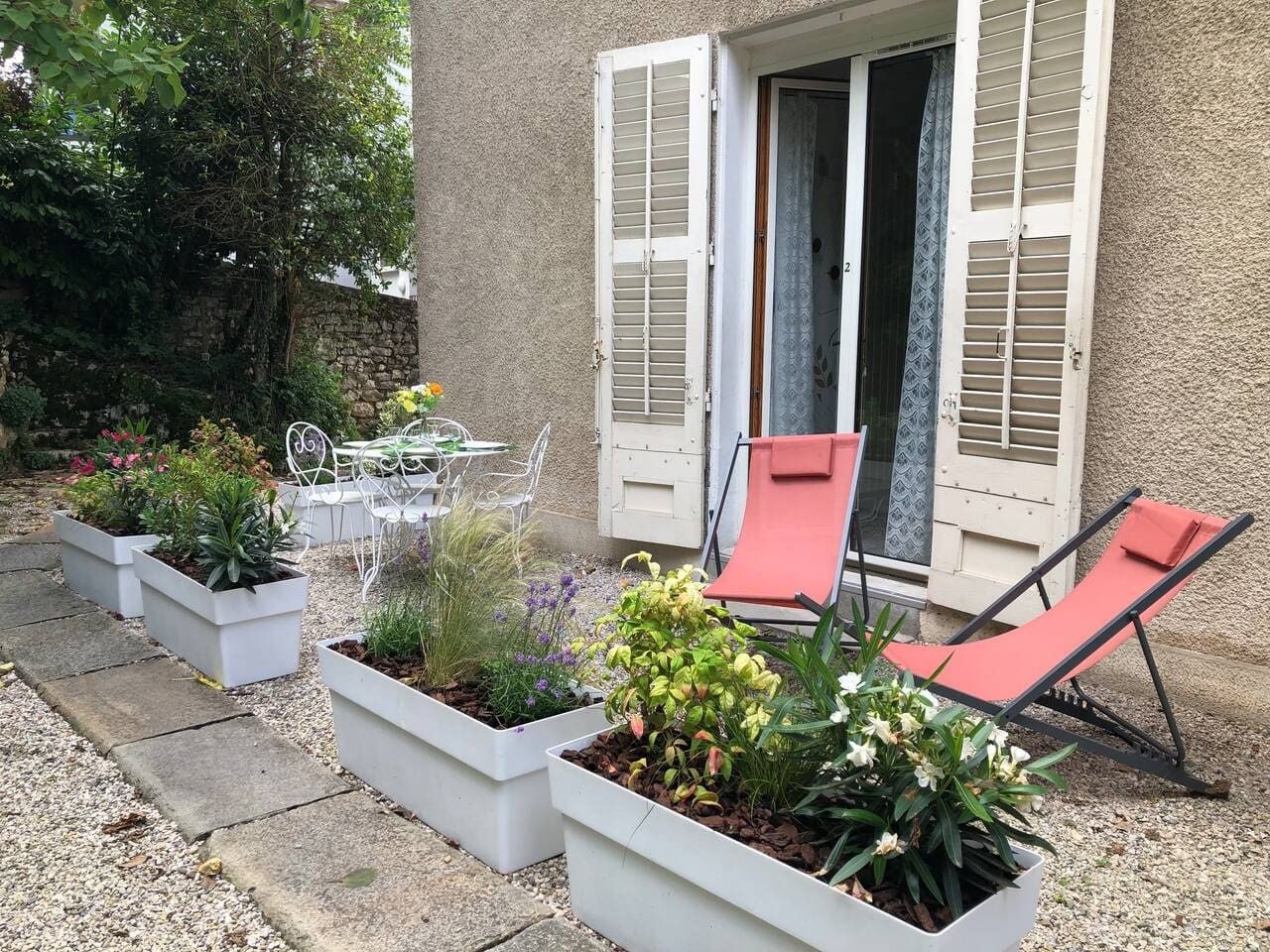 Eine Terrasse mit Gartenmöbeln, Pflanzgefäßen mit verschiedenen Pflanzen und ein Gebäude mit Fensterläden.