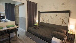 A compact hotel room with a bed, a small desk, a chair, a kitchenette, and a shower area.