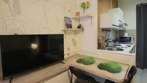 A modern kitchen with a flat-screen TV, a dining table with green leaf-shaped placemats, and a small plant on a shelf.