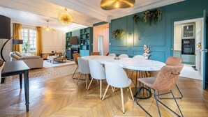 A spacious living room with a large round table, comfortable seating, and a kitchen area visible through an open door.