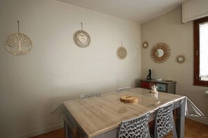 A dining area with a wooden table, white chairs, and decorative wall art including a tree, a heart, and sunburst patterns. - Appartement 55m², 1 chambre, centre-ville, garage (Aix-les-Bains)