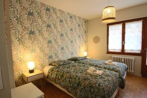 A bedroom with two beds, floral wallpaper, a bedside lamp, a radiator, and a window with curtains. - Appartement 55m², 1 chambre, centre-ville, garage (Aix-les-Bains)