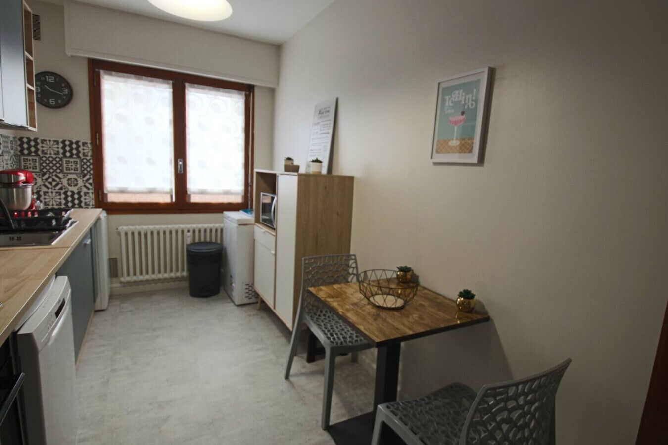 Una cucina funzionale con un tavolo di legno, sedie, un termosifone, un cestino e una sveglia appesa al muro.