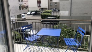 Ein Balkon mit zwei blauen Stühlen und einem Tisch, mit Blick auf eine Straße mit parkenden Autos und Gebäuden.