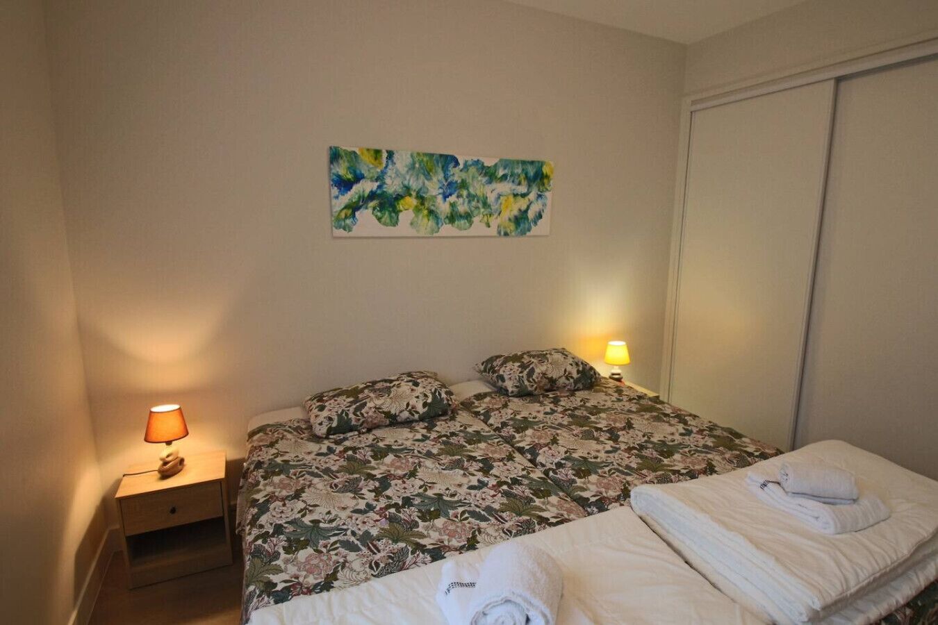 A bedroom with a bed, two bedside lamps, a nightstand, a floral bedspread, and a painting on the wall.