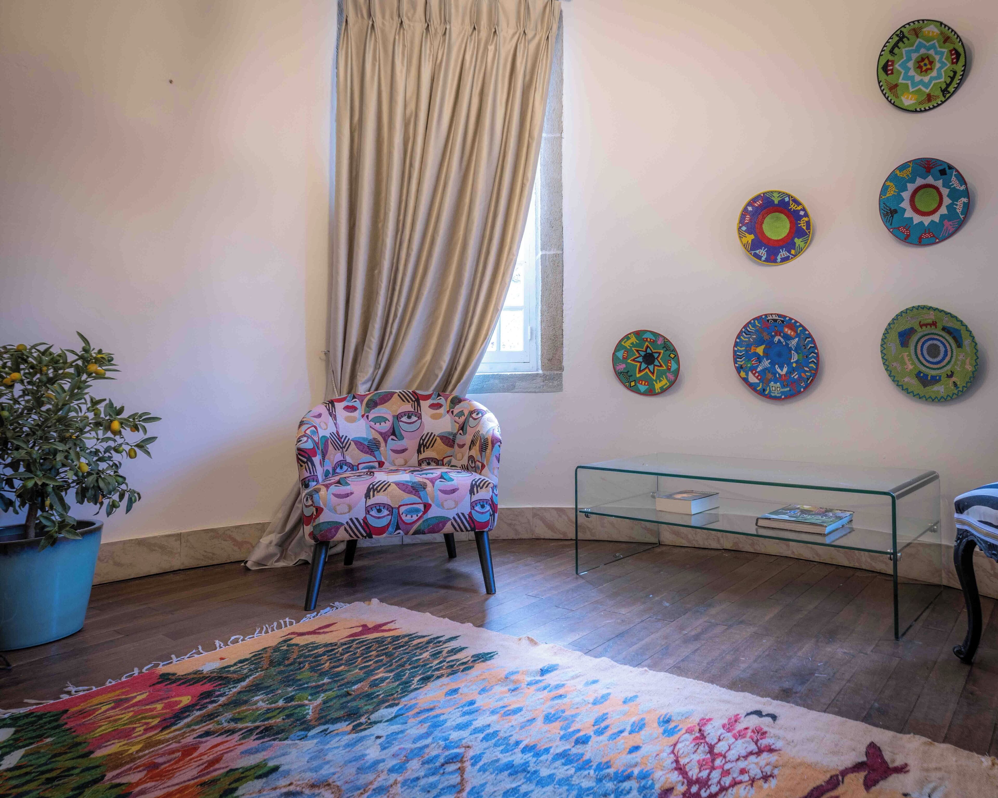 A room with a patterned armchair, a glass coffee table, a colorful rug, a potted plant, and decorative plates on the wall.