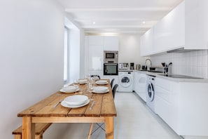 A modern kitchen with white cabinetry, a wooden dining table set for four, a washing machine, and a coffee maker.