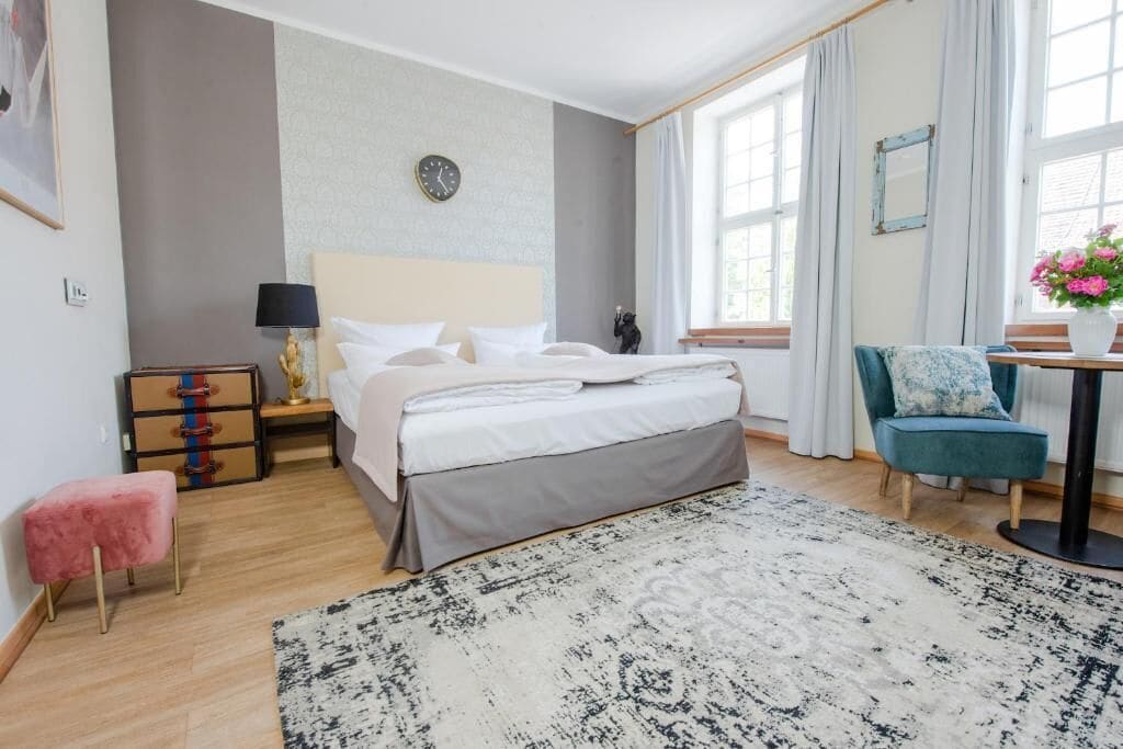 Una habitación de hotel con cama, mesita de noche, lámpara, tocador, silla y vistas al exterior por la ventana.