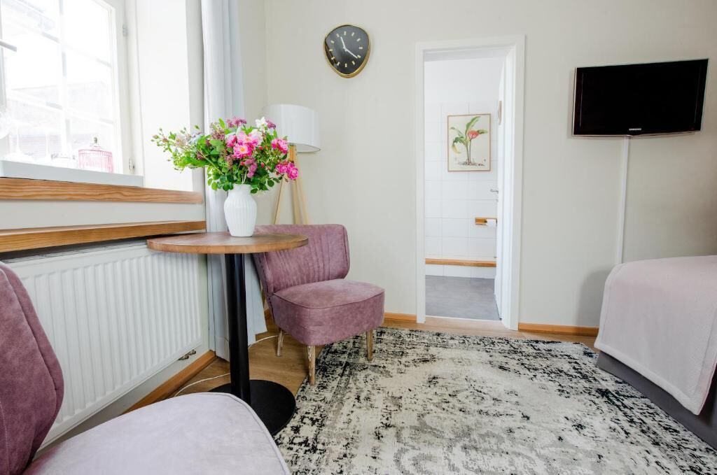 A hotel room with a round table, two pink chairs, a white vase with flowers, a clock, a television, and a patterned rug.