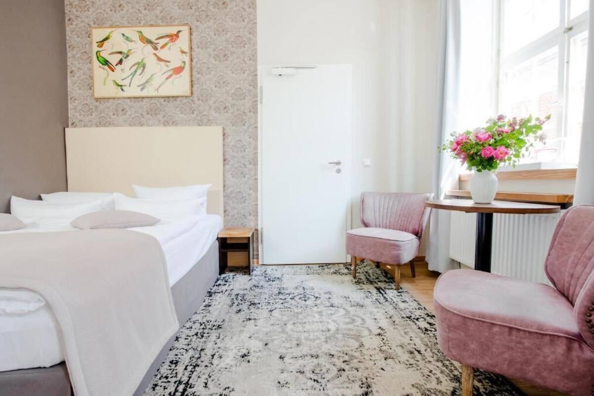 Ein Hotelzimmer mit einem Bett, einem Nachttisch, einem Sessel, einem kleinen Tisch mit Blumenvase und einer Tür.