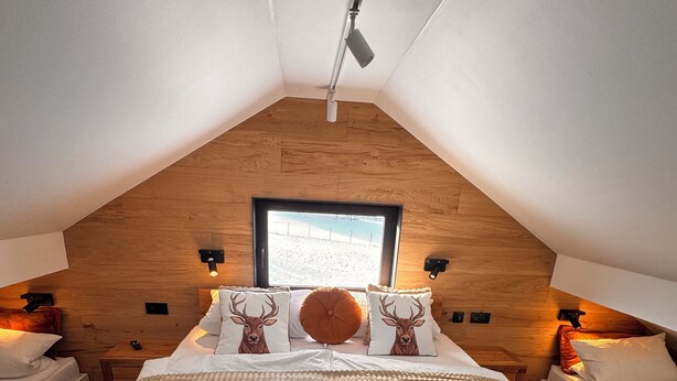 A bedroom with a sloped ceiling, wooden headboard, and a large window. There are two bedside tables with lamps, a bed with a textured blanket, and decorative pillows with deer images. - New vacation home on the outskirts (Eupen)