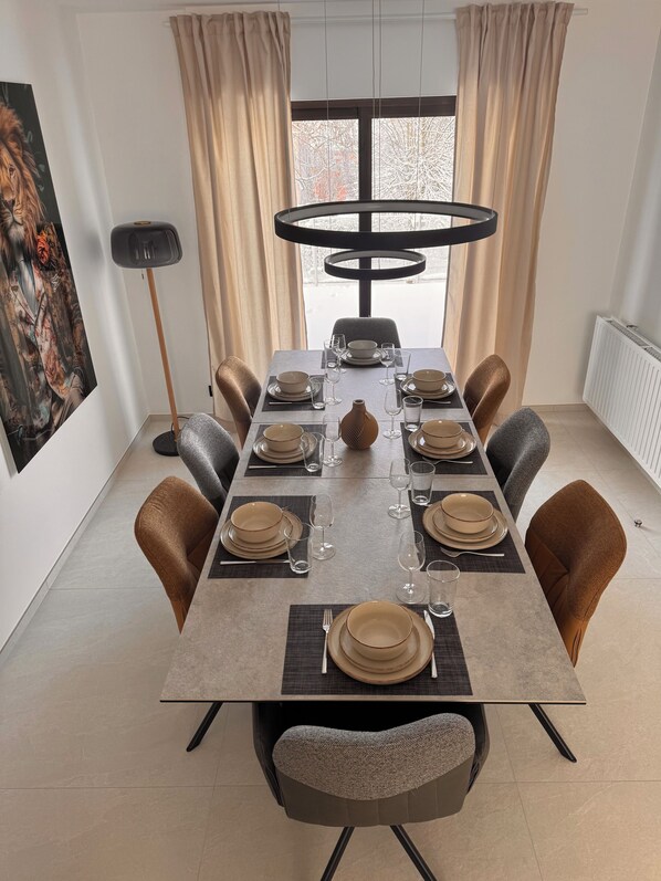 A dining room with a long table set for six, surrounded by six chairs, a modern chandelier, and a large window with curtains. - New vacation home on the outskirts (Eupen)