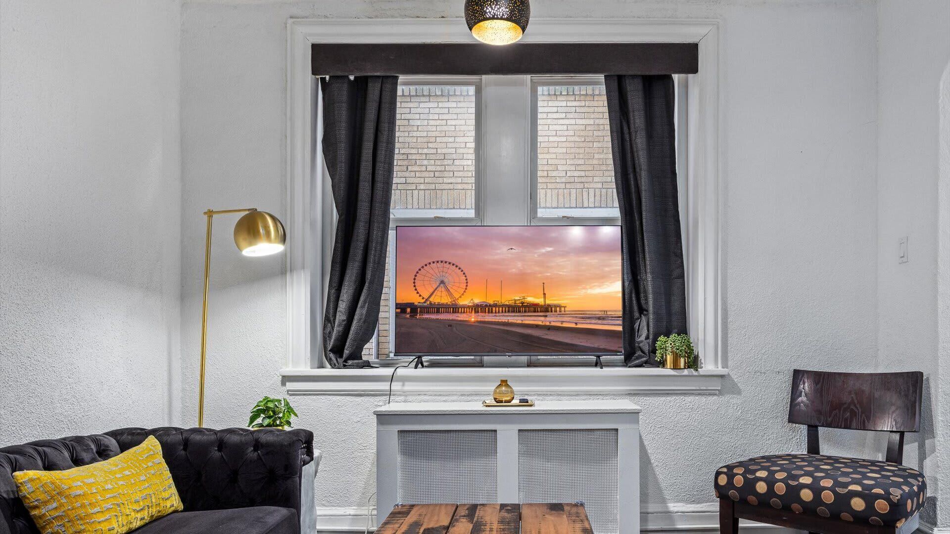 Un salon avec un grand téléviseur fixé à un mur blanc, un canapé noir agrémenté d’un coussin jaune, une table basse en bois et un fauteuil avec un coussin à pois.