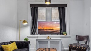 Ett vardagsrum med en stor TV som är monterad på en vit vägg, en svart soffa med en gul kudde, ett träbord och en stol med ett kuddöverdrag med prickar.