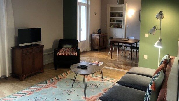 Un soggiorno con divano, tavolino, televisione su mobile, tavolo da pranzo con sedie e angolo cottura con mobiletti e lampada.