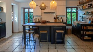 Uma cozinha ampla com uma ilha grande, banquetas, luminárias pendentes e vista para o verde através de amplas janelas.