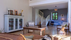Uma sala de estar ampla com pé direito alto, uma escada que dá para um andar superior, uma área de jantar com mesa de madeira e um espaço de estar aconchegante com sofá e poltronas.
