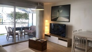 A modern living room with a flat-screen TV, a large window, and a dining area. - Appartement 66m² 2 chambres, garage et terrasse (Aix-les-Bains)