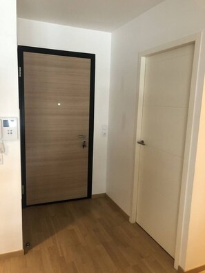 A hallway with two doors, one wooden and one white, and a light switch on the wall. - Appartement 66m² 2 chambres, garage et terrasse (Aix-les-Bains)