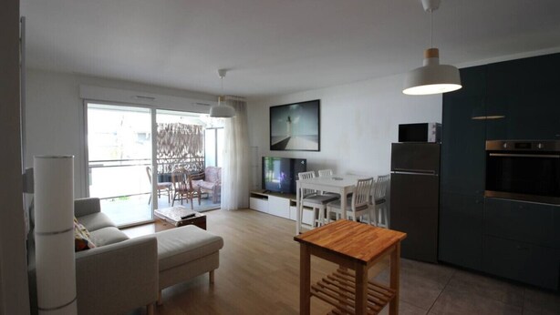 A modern apartment with a living room, dining area, and kitchen. There is a sofa, a wooden table, and a TV mounted on the wall. - Appartement 66m² 2 chambres, garage et terrasse (Aix-les-Bains)
