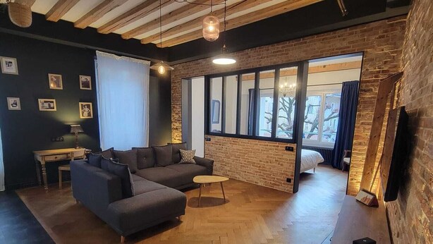 A spacious living area with a herringbone wooden floor, a sectional sofa, a small coffee table, and a large window providing natural light. - Le Loft Des Arts (Thonon-les-Bains)