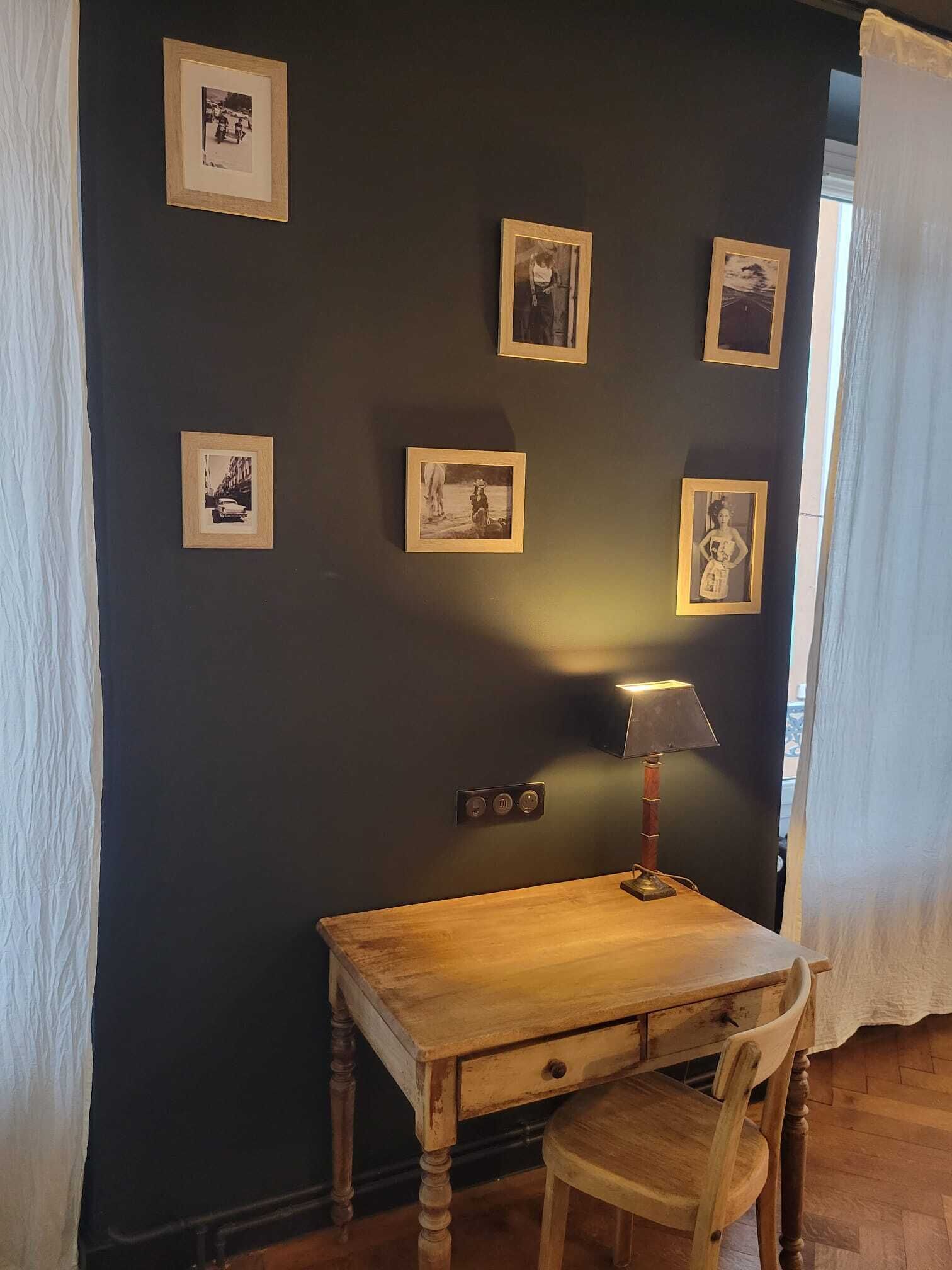 Una habitación con una pared oscura, una mesa de madera, una silla y una lámpara. Hay cuadros en la pared.