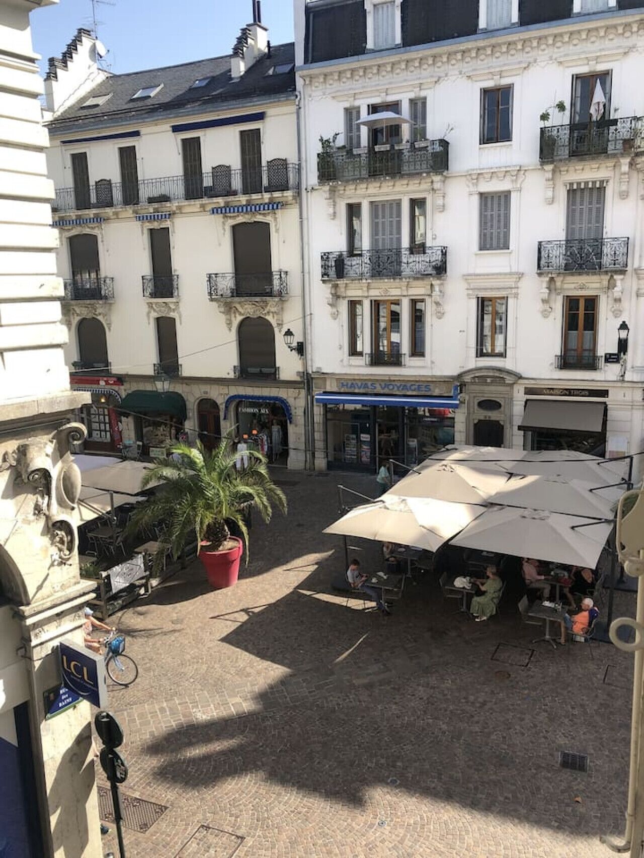 Un patio con mobiliario exterior, una palmera y edificios con balcones y escaparates.