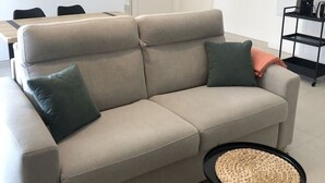 A modern living room with a beige sofa, two green pillows, a woven side table, and a coffee machine on a black shelf. - Appartement 46m² 1 chambre, parking, proche du lac (Aix-les-Bains)