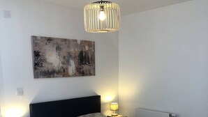A neatly made bed with a floral-patterned bedspread, two bedside lamps, a modern pendant light, and a painting on the wall. - Appartement 46m² 1 chambre, parking, proche du lac (Aix-les-Bains)